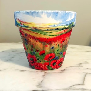 Puede incluir: Maceta de cerámica pintada a mano con una vibrante escena de campo de amapolas. El diseño de la maceta incluye un cielo azul, un paisaje amarillo y verde y numerosas amapolas rojas. El borde está pintado con cielo azul y nubes.