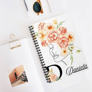 Puede incluir: Un cuaderno encuadernado en espiral blanco con un diseño floral. La portada presenta un dibujo lineal del rostro de una mujer, una gran letra "D" con detalles florales y el nombre "Daniela". Las flores son de color melocotón y amarillo.