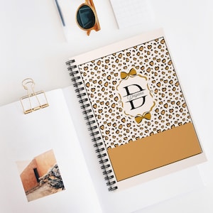Puede incluir: Un cuaderno con espiral con un estampado de leopardo de pequeños corazones marrones sobre un fondo crema. El cuaderno presenta un lazo dorado y un monograma con la letra "D" y el nombre "Dayana". Una sección dorada en la parte inferior.