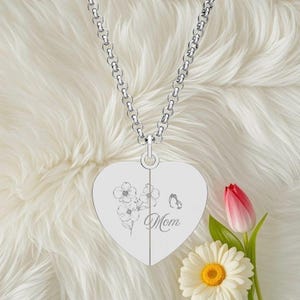 Könnte beinhalten: Silberne herzförmige Anhängerkette mit floralem Design und dem Wort "Mom" eingraviert. Die Kette wird auf einer weißen, flauschigen Oberfläche präsentiert, mit einer rot-rosa Tulpe und einer weißen Gänseblümchen im Hintergrund.