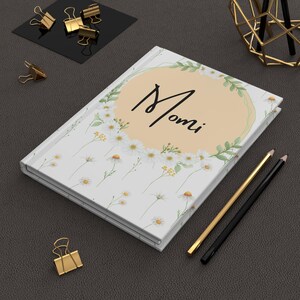 Puede incluir: Un diario blanco con un estampado floral de margaritas y la palabra "Momi" en escritura negra. También se ven lápices dorados y negros, clips dorados y un portalápices geométrico dorado.