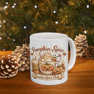 Puede incluir: Una taza de cerámica blanca con una ilustración colorida de bebidas de calabaza, calabazas de Halloween y un pequeño fantasma. El texto "Pumpkin Spice" aparece en la parte superior y "Pumpkin Spice Club" está escrito en una cinta en la base.