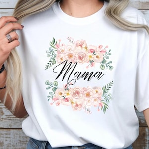 Puede incluir: Camiseta blanca con la palabra "Mama" en elegante escritura negra, rodeada de un arreglo floral de rosas rosas y hojas verdes. El diseño está centrado en la camiseta.
