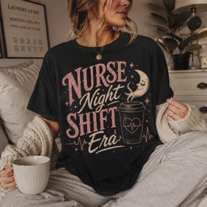 Puede incluir: Camiseta negra con la frase "Nurse Night Shift Era" en rosa y blanco. El diseño incluye una taza de café con un corazón y una luna creciente. La camiseta la lleva una persona.