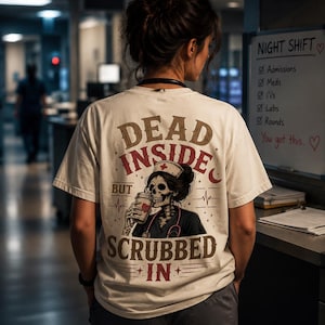 Puede incluir: Camiseta color crema con las palabras "DEAD INSIDE BUT SCRUBBED IN" en letras marrones y burdeos. La camiseta presenta un gráfico de una enfermera esqueleto bebiendo de una taza de café. La persona lleva pantalones grises.