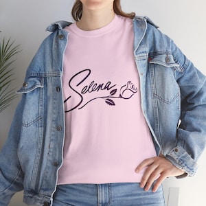 Puede incluir: Camiseta rosa claro con el nombre "Selena" en cursiva, acompañada de una rosa. El diseño es de color púrpura oscuro. La persona lleva una chaqueta vaquera azul.