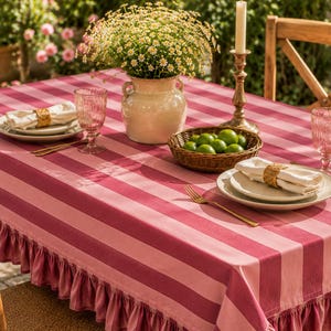 Pode incluir: Uma mesa posta para jantar com uma toalha de mesa listrada rosa e branca. A mesa está posta com pratos, guardanapos, copos e talheres dourados. Um vaso de flores e uma cesta de limas também estão na mesa.