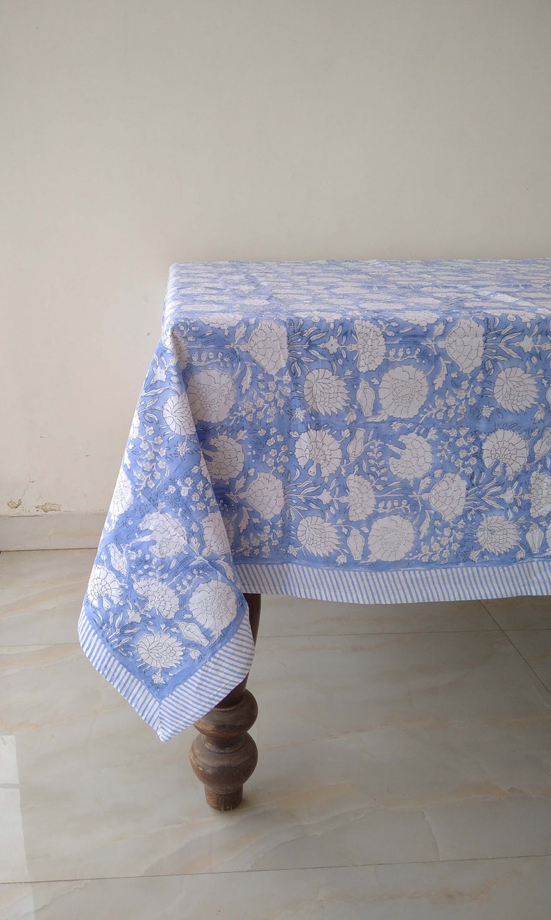 May include: A blue and white floral tablecloth with a striped border. The tablecloth is draped over a wooden table with turned legs.