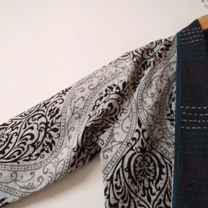 K&ouml;nnte beinhalten: Ein lang&auml;rmeliges Kleidungsst&uuml;ck mit einem schwarzen und wei&szlig;en Paisley-Muster auf dem &Auml;rmel und einem dunkelblauen und schwarzen gemusterten Stoff auf der Vorderseite. Das Kleidungsst&uuml;ck ist gefaltet und h&auml;ngt an einem Kleiderb&uuml;gel.