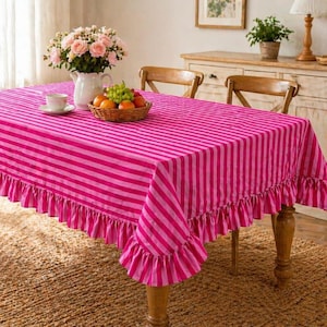 May include: A bright pink and light pink striped tablecloth with a ruffled edge. The tablecloth is on a wooden table with a basket of fruit, a cup and saucer, and a vase of pink roses.