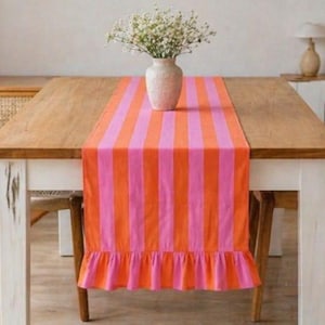 May include: A striped table runner in pink and orange, with a frilled edge. A vase with flowers sits on the runner, which is placed on a wooden table. The table has white legs and is surrounded by wooden chairs. The runner is a decorative item.