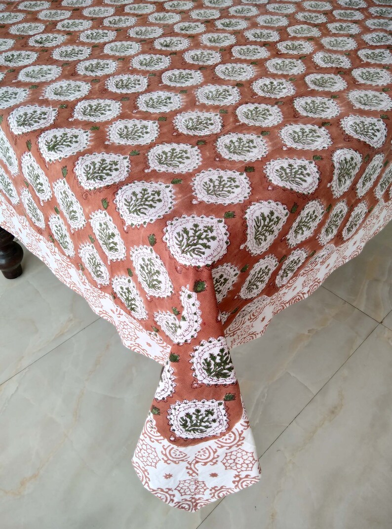 Pu&ograve; includere: Una tovaglia color terracotta con un motivo floreale bianco e verde. La tovaglia ha un bordo a festoni ed &egrave; realizzata in cotone.