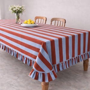 Könnte beinhalten: Rechteckige Tischdecke mit abwechselnd breiten Streifen in Rost und Hellblau. Die Tischdecke hat einen gerüschten Rand und liegt auf einem Holztisch. Eine Vase mit weißen Blumen und ein Teller mit Zitronen stehen auf dem Tisch.
