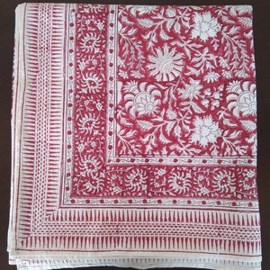 K&ouml;nnte beinhalten: Eine wei&szlig;e Baumwolltischdecke mit rotem Blumendruck. Die Tischdecke hat einen Rand mit einem sich wiederholenden geometrischen Muster.