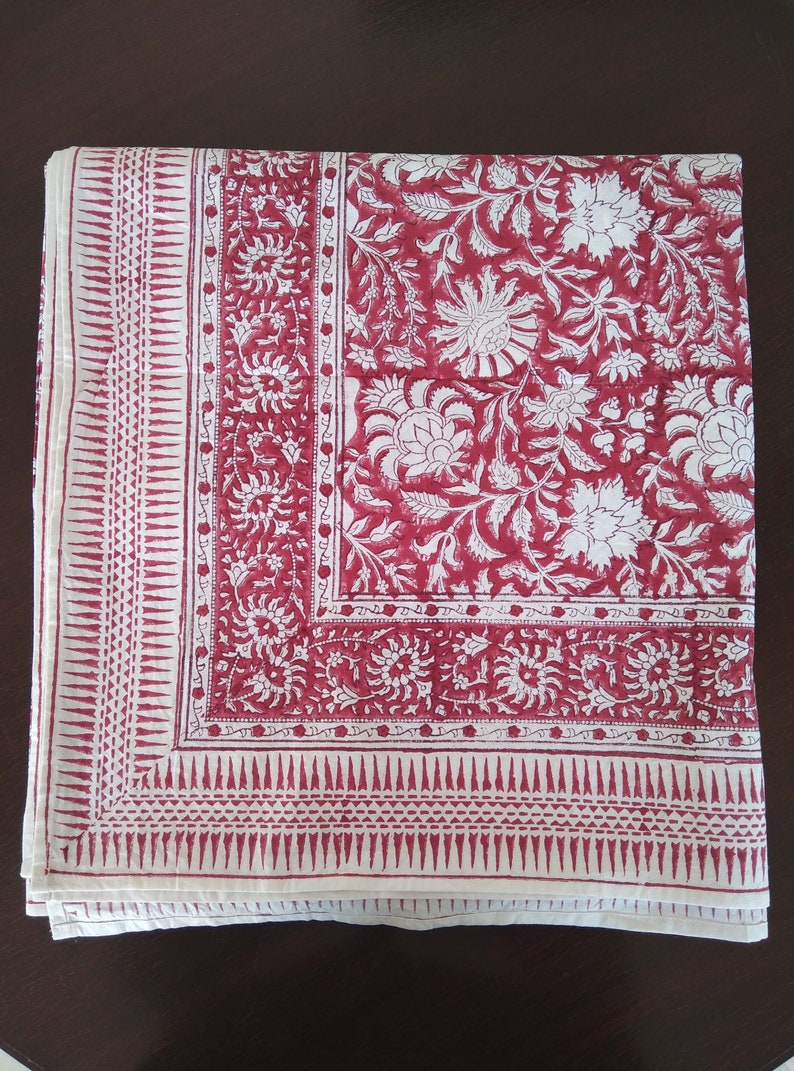 K&ouml;nnte beinhalten: Eine wei&szlig;e Baumwolltischdecke mit rotem Blumendruck. Die Tischdecke hat einen Rand mit einem sich wiederholenden geometrischen Muster.