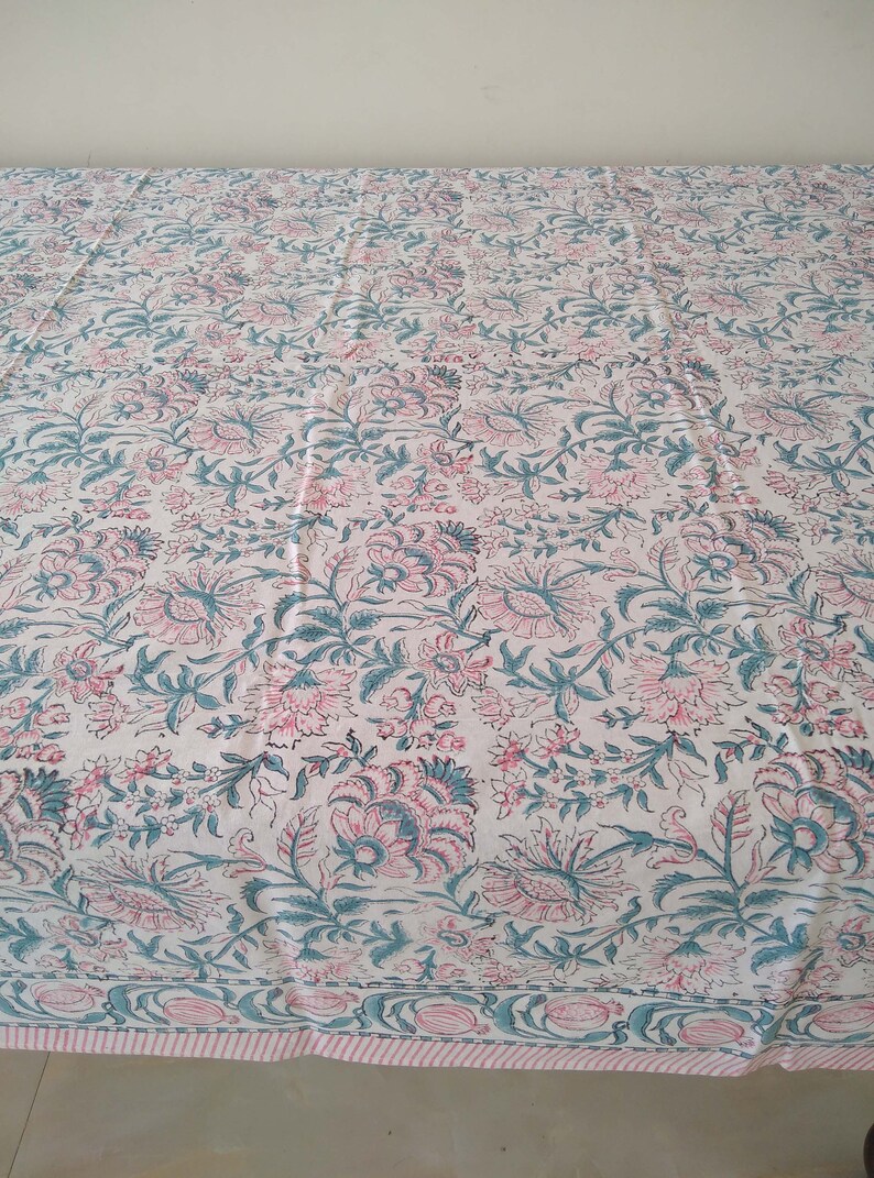 Peut inclure: Une nappe blanche avec un motif floral dans les tons rose et turquoise. La nappe a une bordure avec un motif r&eacute;p&eacute;t&eacute; de rose et de turquoise.