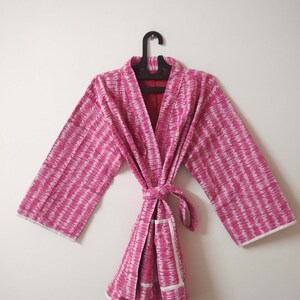 Könnte beinhalten: Ein rosa-weiß gemusterter Bademantel mit weißem Besatz. Der Bademantel hat einen Bindegürtel und eine Tasche auf der linken Seite.