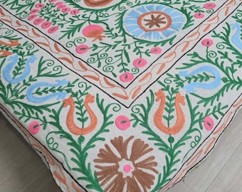 May include: A white linen tablecloth featuring intricate embroidery in shades of green, pink, blue, and brown. The design incorporates floral motifs, swirls, and geometric patterns.