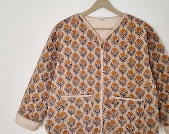 Könnte beinhalten: Beige gesteppte Jacke mit einem Blumenmuster in Gelb und Marineblau. Die Jacke hat einen V-Ausschnitt, zwei Vordertaschen und ist geknöpft. Sie hängt an einem Holzkleiderbügel.