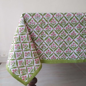 Puede incluir: Un mantel blanco con un patrón floral verde y rosa. El mantel tiene un borde verde y está drapeado sobre una mesa de madera.