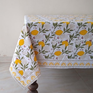 May include: White tablecloth with a lemon print. The tablecloth has a yellow border with a scalloped design. The lemons are yellow with green leaves and red berries.