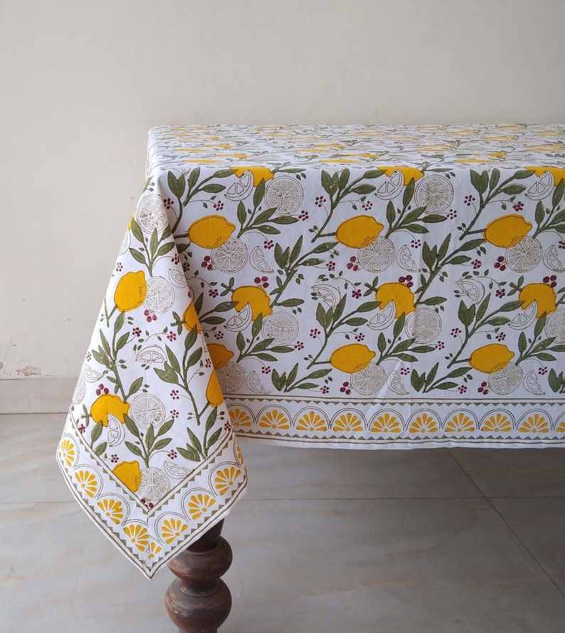 May include: White tablecloth with a lemon print. The tablecloth has a yellow border with a scalloped design. The lemons are yellow with green leaves and red berries.