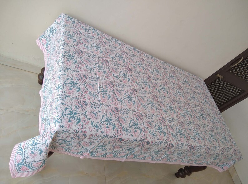 Peut inclure: Une nappe blanche avec un imprim&eacute; floral rose et vert. La nappe a une bordure rose et est drap&eacute; sur une table en bois.