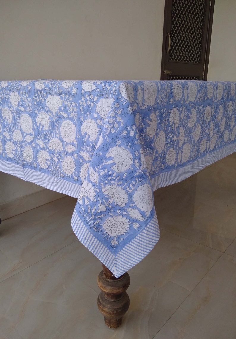 May include: A blue and white floral tablecloth with a striped border. The tablecloth is draped over a wooden table with turned legs.