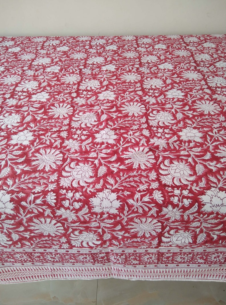 K&ouml;nnte beinhalten: Rotes Baumwoll-Bettlaken mit wei&szlig;em Blumenmuster. Das Laken hat einen Rand mit einem wei&szlig;en und roten geometrischen Muster.