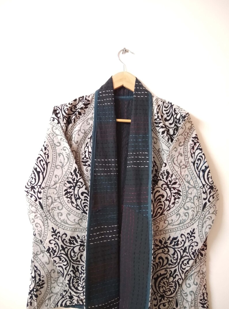 K&ouml;nnte beinhalten: Ein Kimono mit schwarz-wei&szlig;em Muster und einer gesteppten Innenfutter in Tealblau und Schwarz. Der Kimono h&auml;ngt an einem Holzb&uuml;gel.