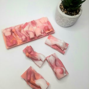 May include: Five pink and white soap bars shaped like human figures. The soap bars are arranged in a row on a white surface.