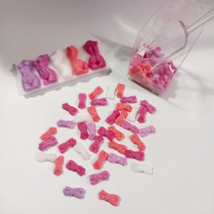 Puede incluir: Una variedad de pequeñas figuras de caramelo rosa, blanco y morado con forma de personas. El caramelo está en un recipiente de plástico transparente con una pala. Parte del caramelo está esparcido sobre una superficie blanca.