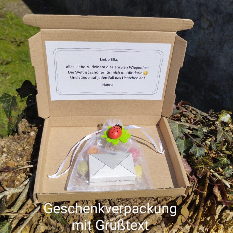 May include: A brown cardboard box with a white card inside. The card has a handwritten message that says "Liebe Ella, alles Liebe zu deinem diesj&auml;hrigen Wiegenfest. Die Welt ist sch&ouml;ner f&uuml;r mich mit dir darin Und z&uuml;nde auf jeden Fall das Lichtchen an! Hanna". The box also contains a small white envelope and a clear bag with colorful candies and a ladybug decoration.