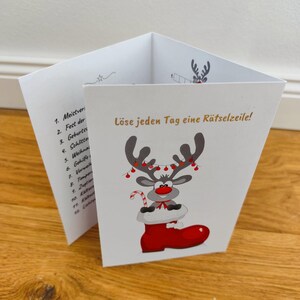 Può includere: Un biglietto di Natale bianco piegato con una renna dei cartoni animati in uno stivale rosso. La renna ha corna decorate con luci rosse e un bastoncino di zucchero. Il testo tedesco "Löse jeden Tag eine Rätselzeile!" è stampato.