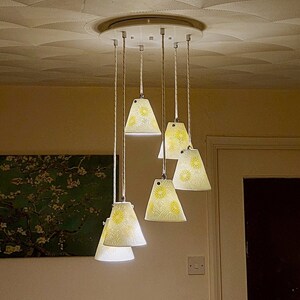May include: A ceiling light fixture with six white lampshades featuring a yellow floral pattern. The lampshades are suspended from a circular metal base.