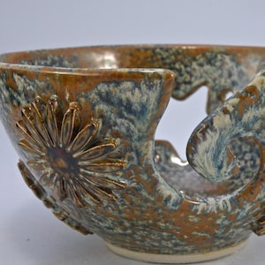 May include: A ceramic yarn bowl with a brown, blue, and cream glaze. The bowl features a daisy design and a curved opening for yarn. The bowl is approximately 6 inches in diameter.