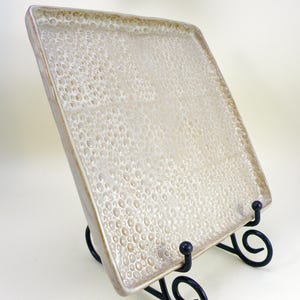 May include: A rectangular ceramic serving tray with a textured, bubbly surface in shades of cream and beige. The tray has a slightly raised edge and is displayed on a black metal stand. The tray's design gives it a unique, handcrafted appearance.
