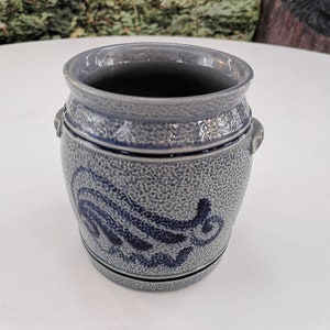 Könnte beinhalten: Eine kleine, graue Keramikdose mit blau-weißem Design. Die Dose hat zwei kleine Griffe an den Seiten und eine strukturierte Oberfläche.