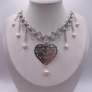 Puede incluir: Collar plateado con un gran colgante calado en forma de corazón. El collar presenta un diseño de cadena con cadenas colgantes, pequeñas perlas y detalles de cristal transparente. El collar se exhibe en un maniquí blanco.