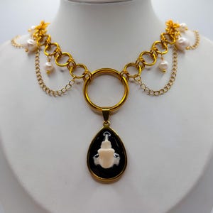 May include: A gold-toned necklace featuring a central teardrop pendant with a black background and a white anatomical bone-like figure. The chain consists of large circular links, delicate gold chains, and small white pearls at the sides.
