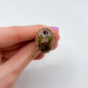Puede incluir: Un anillo de plata con una piedra ovalada transparente que presenta un diseño de flor prensada. La flor es marrón y verde con un centro blanco.