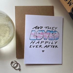 May include: A white card with the text "And they loved happily ever after" in black marker. The word "loved" is written in large, colorful letters with a watercolor effect in shades of blue, pink, and white.