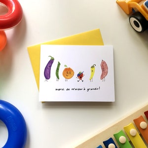 May include: A white card with a colorful illustration of vegetables and a small person. The text on the card reads "merci de m'aider &agrave; grandir!" which translates to "thank you for helping me grow!"