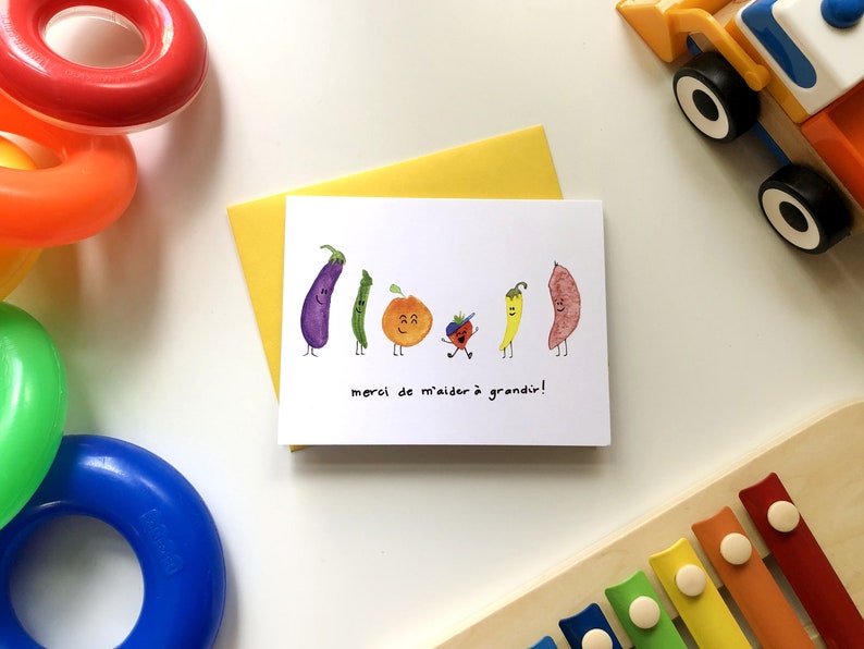 May include: A white card with a colorful illustration of vegetables and a small person. The text on the card reads "merci de m'aider &agrave; grandir!" which translates to "thank you for helping me grow!"
