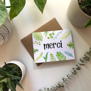 May include: A white card with a green watercolor floral design and the word "merci" in black lettering. The card is sitting on a brown envelope.