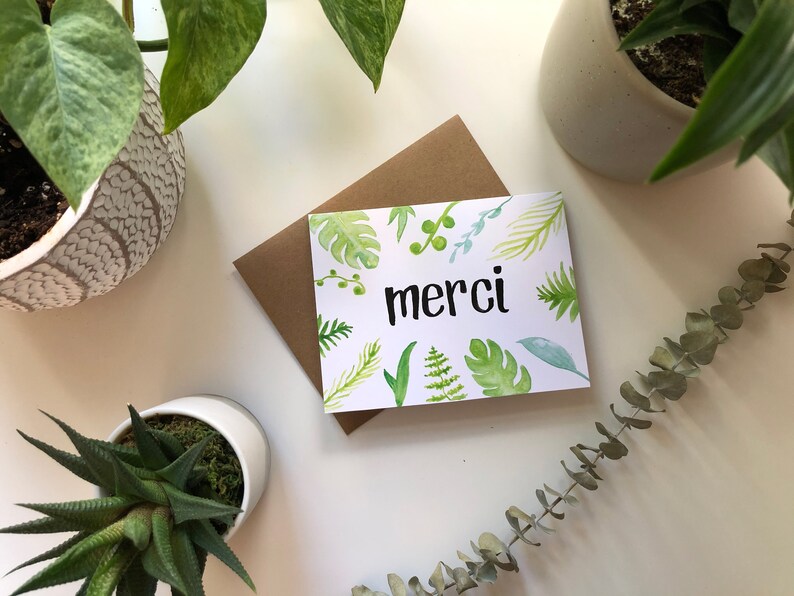 May include: A white card with a green watercolor floral design and the word "merci" in black lettering. The card is sitting on a brown envelope.