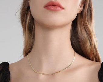 Könnte beinhalten: Eine goldene Kettenkette mit einem schlichten, flachen Design. Die Kette wird von einer Person mit langen, blonden Haaren getragen.