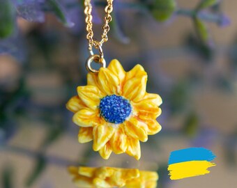 May include: A gold chain necklace with a yellow sunflower pendant. The sunflower has a blue center and is surrounded by green leaves. The necklace is hanging from a purple plant.