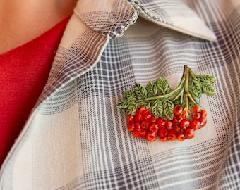 May include: A ceramic brooch shaped like a cluster of bright red rowan berries with textured green leaves. The piece features a glossy finish on the berries and detailed veining on the foliage, attached to a plaid button-down shirt with a white button.