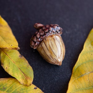 Op de afbeelding: Een keramische eikel met een bruine en witte glazuur, liggend op een zwart oppervlak met twee gele bladeren.